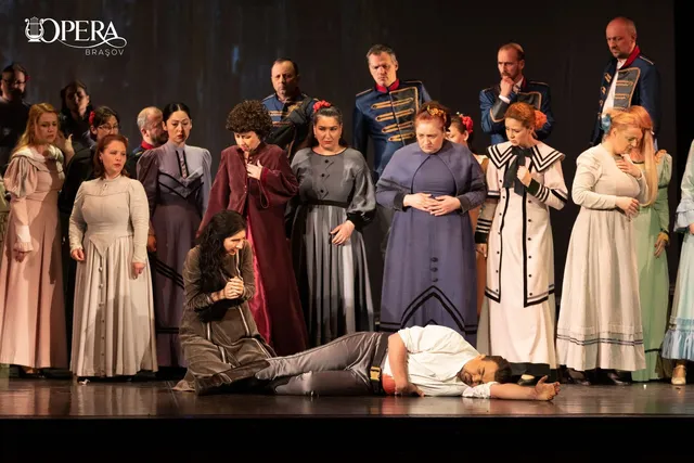 Un grup de actori în costume de epocă stă pe o scenă întunecată, în timp ce un bărbat este întins pe podea în prim-plan.