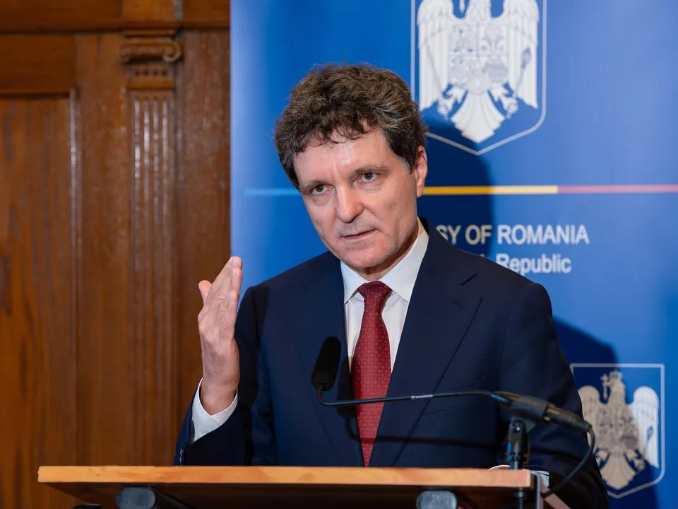 Un bărbat cu părul închis la culoare, purtând un costum închis la culoare, cămașă albă și cravată roșie, stă în spatele unui podium cu microfon.
