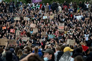 O mulțime mare de oameni ridică mâinile și țin diverse panouri de protest într-un spațiu deschis.