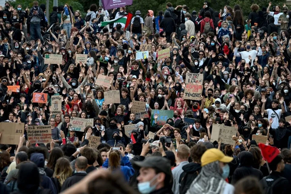 O mulțime mare de oameni ridică mâinile și țin diverse panouri de protest într-un spațiu deschis.