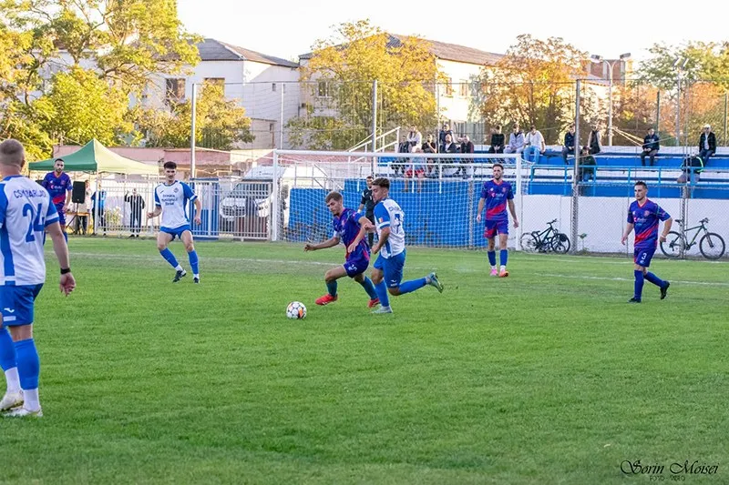 Câțiva jucători de fotbal în echipamente albastre și violet se află pe un teren de gazon verde în timpul unui meci.