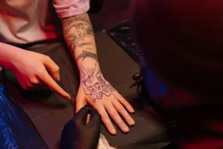 O persoană cu mănuși negre tatuază mâna unei alte persoane care are un tatuaj pe antebraț și pe dosul palmei.