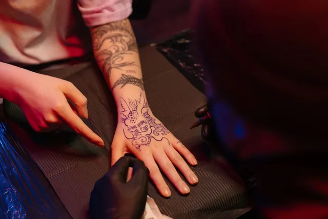 O persoană cu mănuși negre tatuază mâna unei alte persoane care are un tatuaj pe antebraț și pe dosul palmei.