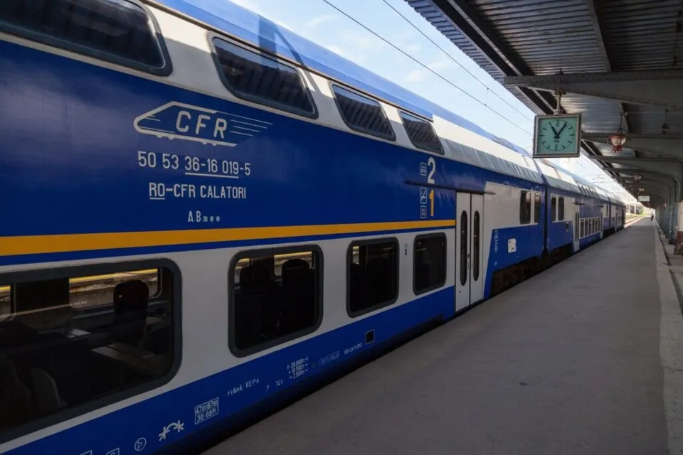 Un tren de pasageri albastru și alb cu ferestre mari stă oprit pe un peron de gară lângă un ceas suspendat.