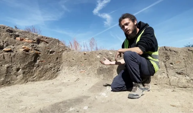 Un bărbat cu barbă, purtând hanorac negru și vestă reflectorizantă, stă ghemuit pe un teren de excavare lângă un zid de pământ și piatră.