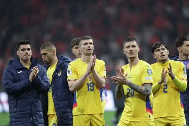 Câțiva bărbați îmbrăcați în echipamente sportive galbene și jachete albastre aplaudă pe un teren de fotbal în fața unui public numeros.