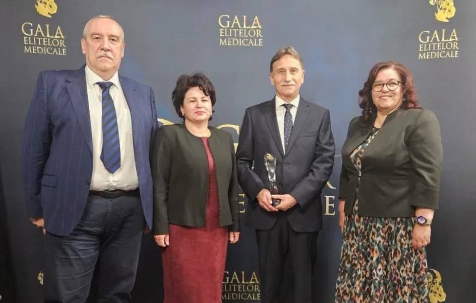 Patru persoane îmbrăcate în haine formale stau în picioare în fața unui fundal cu inscripții aurii.