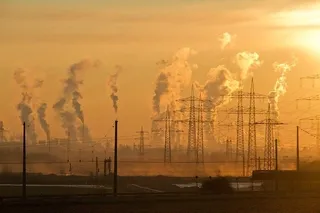 Turnuri de înaltă tensiune și coloane de fum se ridică deasupra unui peisaj industrial sub un cer portocaliu la apus.