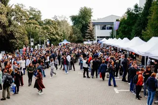O mulțime de oameni stă pe o esplanadă pavată între copaci, corturi albe și o clădire cu ferestre mari.