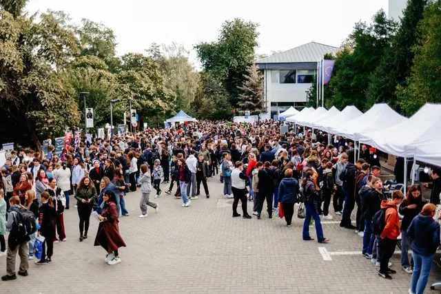 O mulțime de oameni stă pe o esplanadă pavată între copaci, corturi albe și o clădire cu ferestre mari.