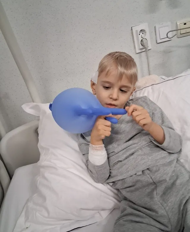 Un copil cu părul blond stă în pat și ține un balon albastru cu ambele mâini.
