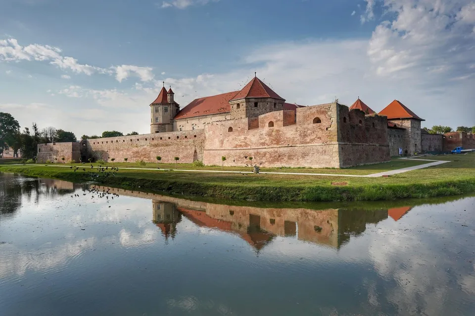 O cetate cu ziduri de cărămidă și turnuri cu acoperișuri în pini este reflectată într-un lac aflat în prim-plan sub un cer înnorat.