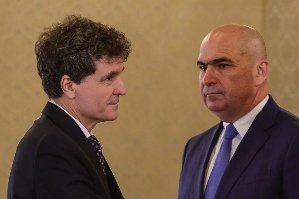 Un bărbat cu părul șaten și creț privește un bărbat chel, îmbrăcat în costum și cravată, într-un decor cu un perete de culoare neutră.