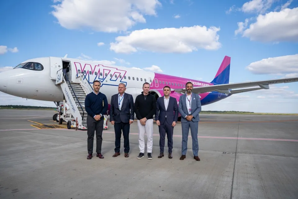 Cinci bărbați stau în rând pe o pistă de beton în fața unui avion alb cu accente roz și albastru sub un cer cu nori.