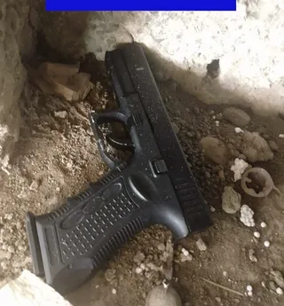 O pistolă neagră este așezată pe un sol nisipos și pietros lângă un perete alb.