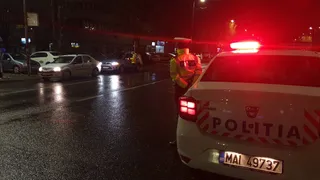 Un polițist în uniformă portocalie stă lângă partea din spate a unei mașini albe marcate cu inscripția POLITIA pe un drum umed noaptea.