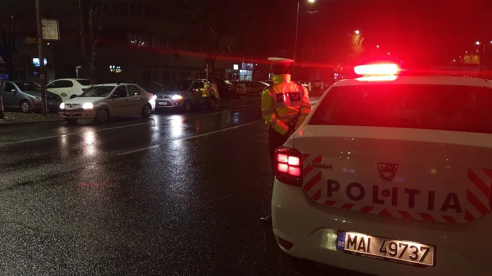 Un polițist în uniformă portocalie stă lângă partea din spate a unei mașini albe marcate cu inscripția POLITIA pe un drum umed noaptea.