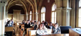 Un grup de persoane stă la bănci în interiorul unei săli cu ferestre înalte și arcade de sticlă, privind spre partea din față a camerei.