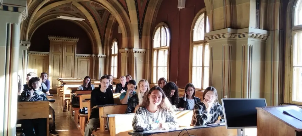 Un grup de persoane stă la bănci în interiorul unei săli cu ferestre înalte și arcade de sticlă, privind spre partea din față a camerei.
