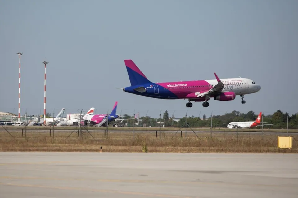 Un avion de pasageri de culoare roz și albă zboară deasupra unei piste de aterizare, având alte aeronave parcate în fundal.