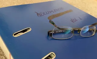 Ochelari cu ramă neagră sunt așezați pe o dosar albastru pe care este scris cu marker negru textul DOCUMENTE VOL. 1.