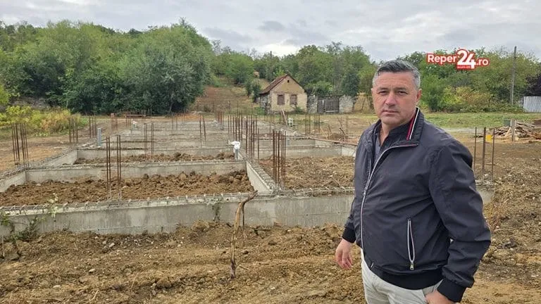 Un bărbat stă în picioare în fața unei fundații de beton de pe un teren de construcție, sub un cer înnorat.