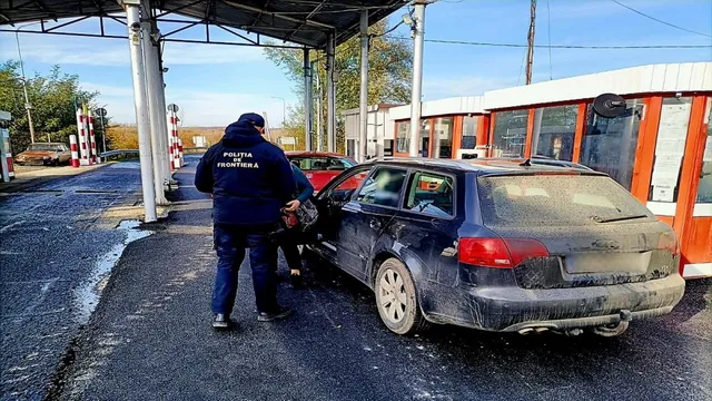 Un agent de poliție stă lângă o mașină neagră de tip break parcată pe un drum asfaltat lângă o clădire cu ferestre mari.