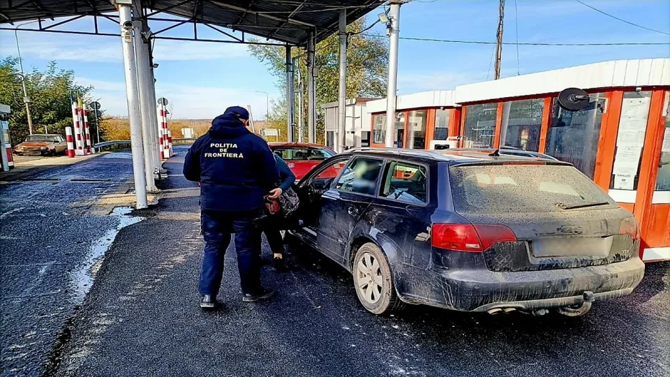 Un agent de poliție stă lângă o mașină neagră de tip break parcată pe un drum asfaltat lângă o clădire cu ferestre mari.