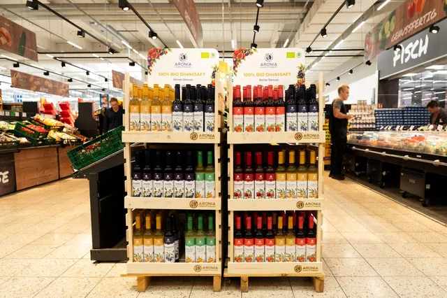 Mai multe rafturi de lemn conțin sticle de suc colorate, dispuse în rânduri pe trei niveluri într-un magazin alimentar.
