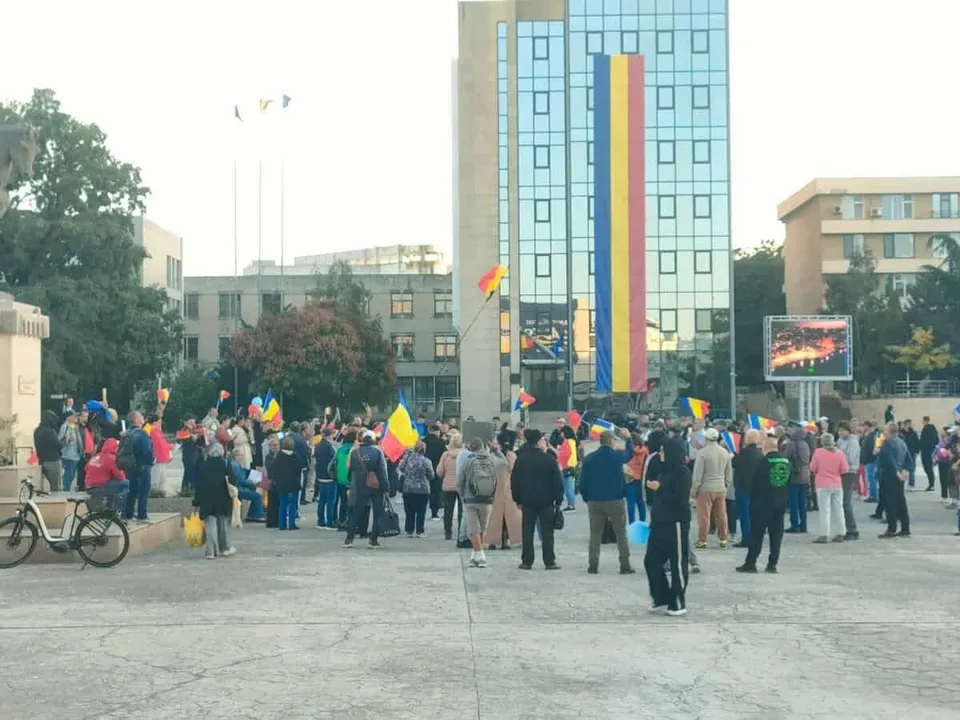 Un grup de persoane stă adunat pe o piață pavată în fața unei clădiri înalte cu fațadă de sticlă și un steag vertical lung.