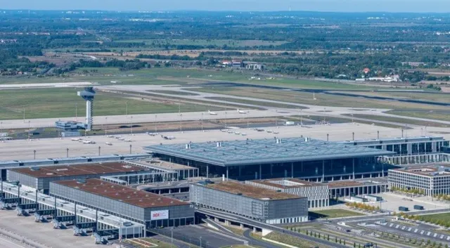 O vedere aeriană a unui terminal de aeroport cu piste, clădiri de terminal și vegetație în fundal sub un cer senin.