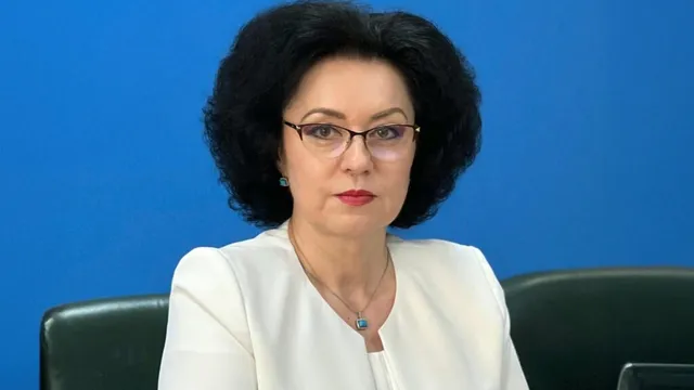 O femeie cu părul negru, purtând ochelari și o bluză albă, stă în fața unui fundal albastru.