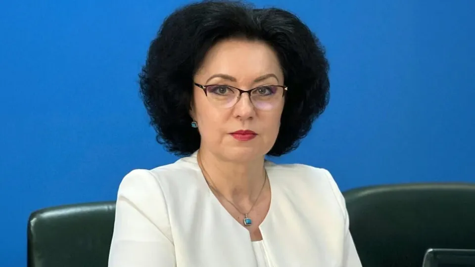 O femeie cu părul negru, purtând ochelari și o bluză albă, stă în fața unui fundal albastru.
