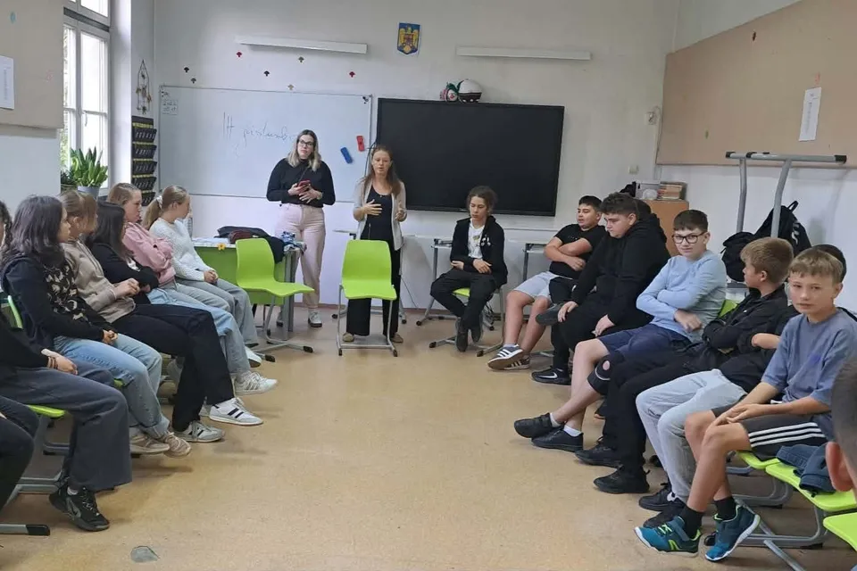 Un grup de elevi stă pe scaune dispuse în cerc într-o sală de clasă, în timp ce două femei stau în picioare în fața lor.