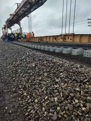 Oamenii în veste reflectorizante stau lângă o structură metalică de culoare galbenă și șine de tren deasupra unui drum de pietriș.