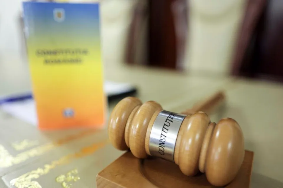 Un ciocan de lemn cu un inel metalic pe care scrie CONSTITUTION se află pe o suprafață de lemn lângă un document colorat.