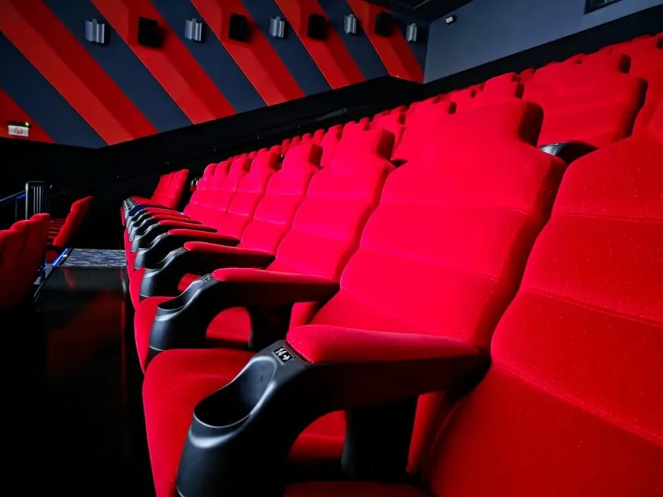 Rânduri de scaune de cinema din catifea roșie cu accesorii negre sunt dispuse în interiorul unei săli întunecate.