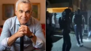 Un bărbat cu părul cărunt stă la o masă, iar în partea dreaptă se observă siluete întunecate de persoane pe un drum noaptea.
