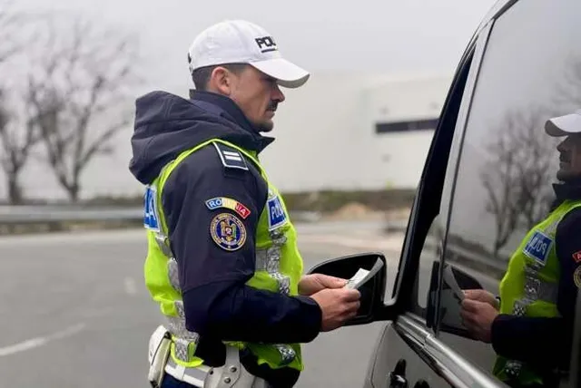 Un polițist în uniformă cu vestă reflectorizantă ține un document lângă fereastra deschisă a unei mașini gri.