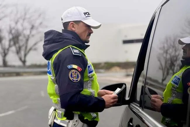 Un polițist în uniformă cu vestă reflectorizantă ține un document lângă fereastra deschisă a unei mașini gri.