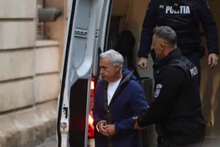 Un bărbat în vârstă, cu părul gri, în costum albastru și cu mainsile legate, este însoțit de doi polițiști lângă o mașină poliție.