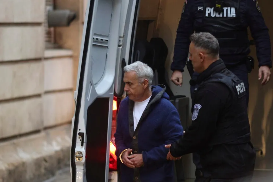 Un bărbat în vârstă, cu părul gri, în costum albastru și cu mainsile legate, este însoțit de doi polițiști lângă o mașină poliție.