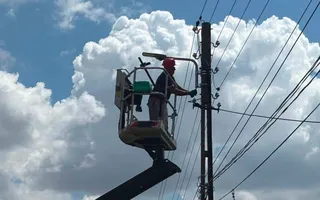 Un muncitor cu cască roșie lucrează într-un coș suspendat de o platformă hidraulică lângă un stâlp electric cu mai multe cabluri sub un cer înnorat.