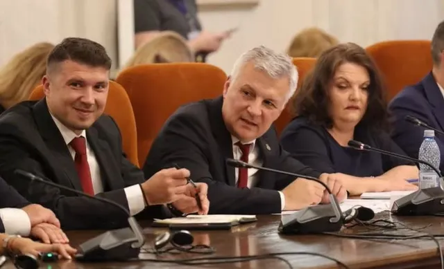 Trei persoane stau la o masă de conferințe cu microfoane, un bărbat din centru purtând costum și cravată roșie, vorbind către cineva.