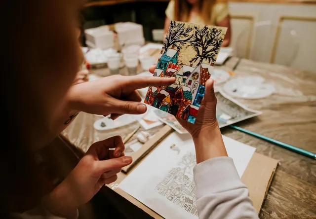 O persoană ține o pictură colorată cu case și un copac, în timp ce degetul celeilalte persoane indică spre desenul respectiv pe o masă de lucru.