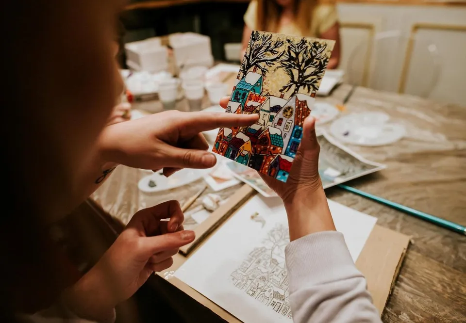 O persoană ține o pictură colorată cu case și un copac, în timp ce degetul celeilalte persoane indică spre desenul respectiv pe o masă de lucru.