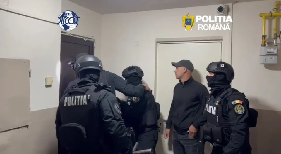 Polițiști echipați cu echipamente tactice negre și cască sunt într-o cameră, observând un bărbat în picioare lângă o ușă.