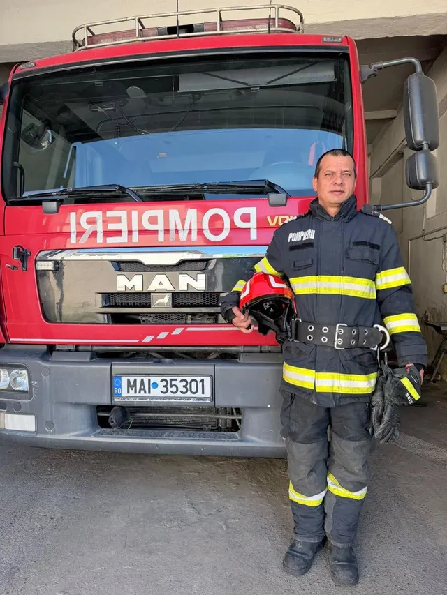 Un bărbat în echipament de pompier stă în picioare în fața unui camion roșu cu inscripția POMPIERI și plăcuță de înmatriculare MAI 35301.