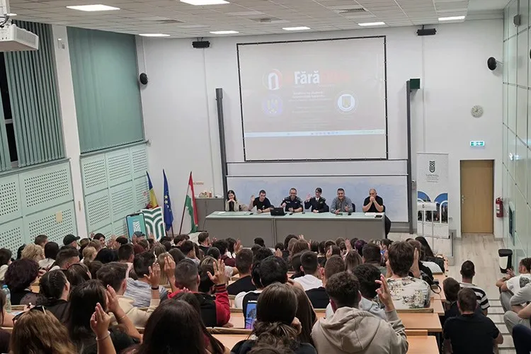 Un grup de persoane stă la mese în fața unui ecran de proiecție, în timp ce un public stă pe scaune în fața lor într-o sală de conferințe.