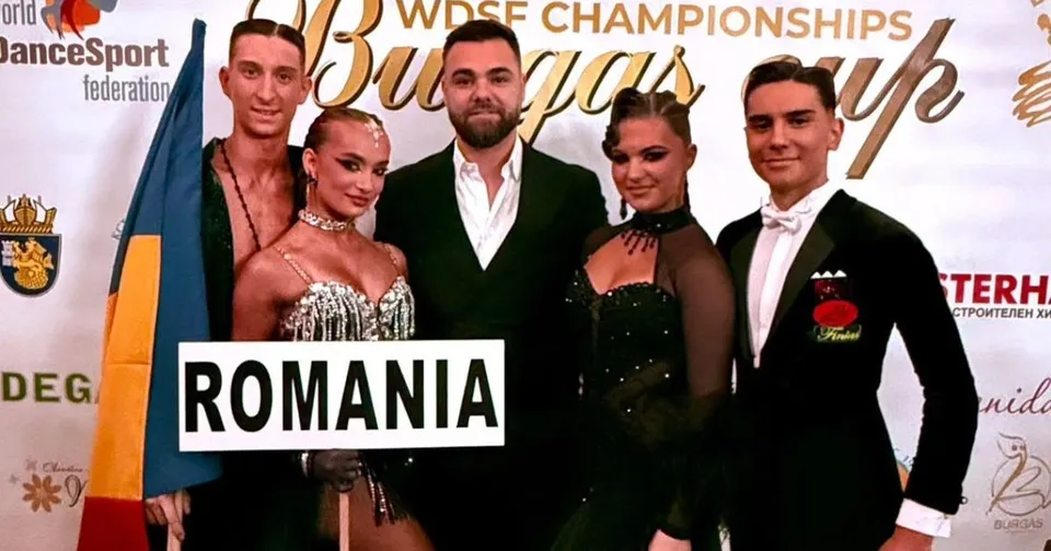 Patru persoane stau în picioare în fața unui panou alb, două femei purtând rochii de seară și doi bărbați în costume elegante.